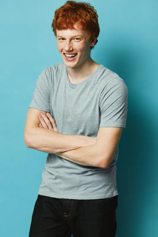 Young man with red hair smiling confidently standing with arms crossed against blue background in casual gray t shirt and black pants.