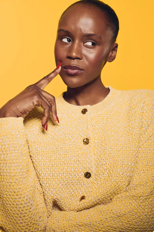 Thoughtful dark skinned African American woman in yellow knitted sweater poses with finger on cheek, looking sideways on bright yellow background. Concept for contemplation and fashion.