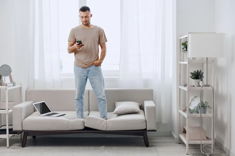 Young man standing on a couch, looking at his smartphone with a thoughtful expression. Stylish modern living room with bright natural light and minimal decor.