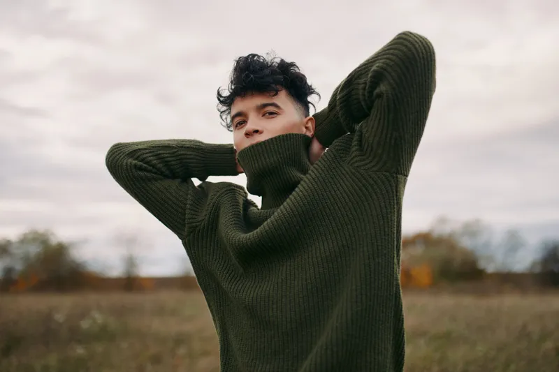 A candid portrait of a young man in a thick green sweater, exuding authenticity and quiet confidence under soft outdoor light, with a calm, natural expression and subtle mood.