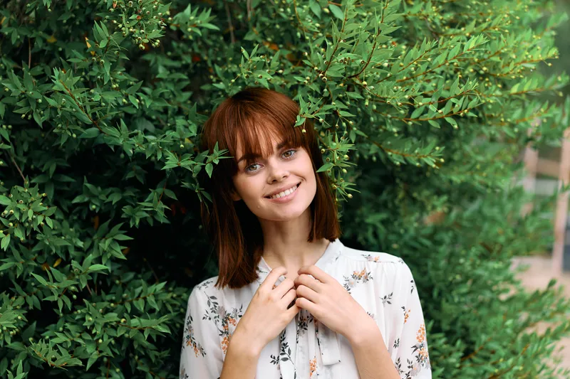 Яркая рыжеволосая женщина стоит на фоне пышного зелёного куста на природе