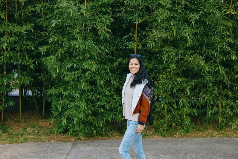Smiling woman walks along a paved walkway beside lush green hedges, wearing a colorblock jacket and light jeans, casual outdoor scene capturing approachable style and urban nature contrast