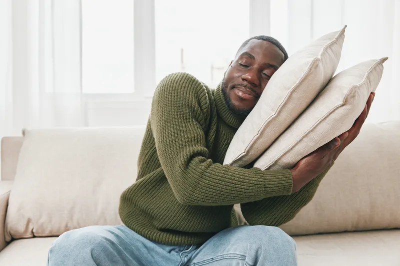 A relaxed man of African descent in a cozy sweater enjoys a moment of comfort by hugging soft pillows, evoking feelings of warmth and tranquility in a light filled room