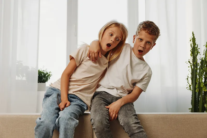 Two surprised children sit on a couch with arms around each other in a bright room. Concept of childhood, friendship, and playful emotions.