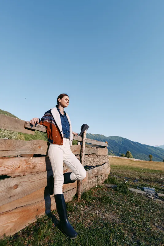 Woman leaning on wooden fence in mountains countryside meadow under clear blue sky, wearing jacket and boots, full body portrait in rural outdoors setting for fashion and travel.