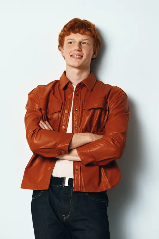 Young man with red hair standing with arms crossed wearing brown leather jacket and jeans, smiling confidently against white background.