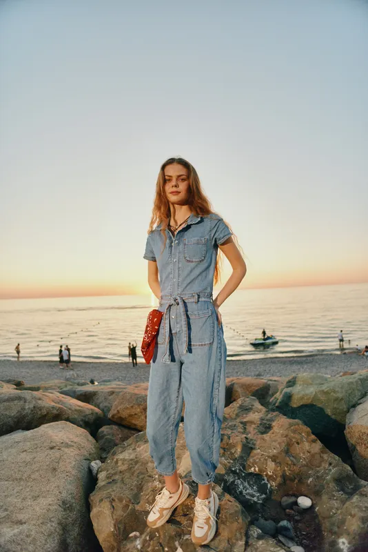 Lifestyle woman in modern boho-western denim street style stands on rocky beach at sunset with film color tones, casual outfit, relaxed mood, ocean background.