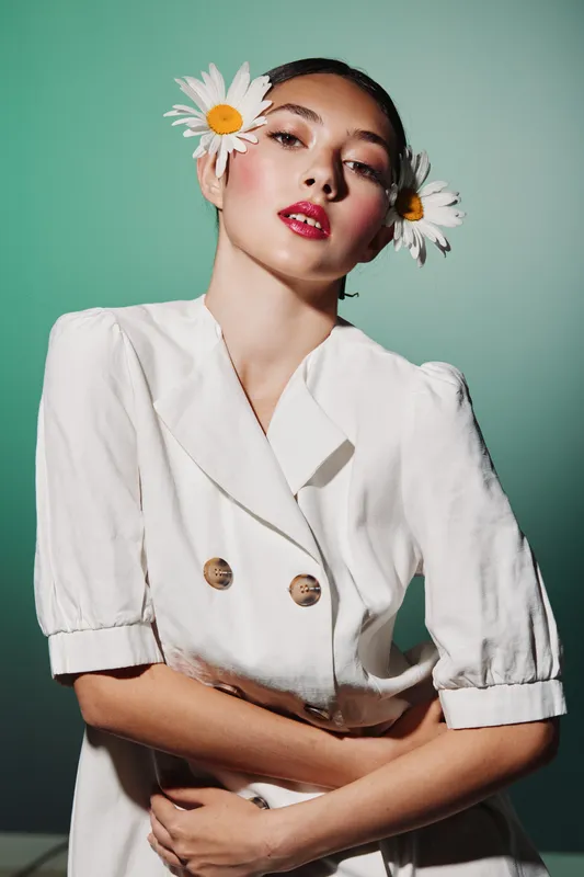 Fashion model with vibrant makeup and big white flowers in hair poses emotionally in white puff sleeve dress. Beauty concept with natural decor background and elegant styling.