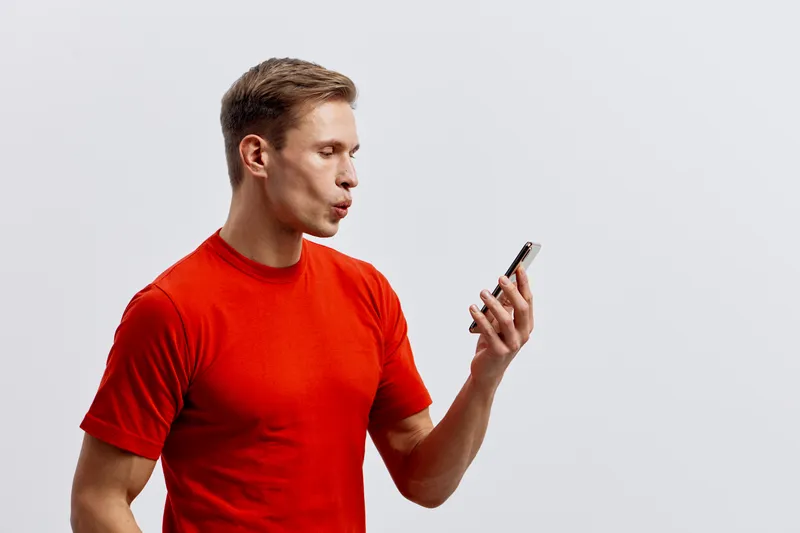Young Caucasian man in red t-shirt looking at mobile phone with focused expression on plain white background studio portrait. People technology concept