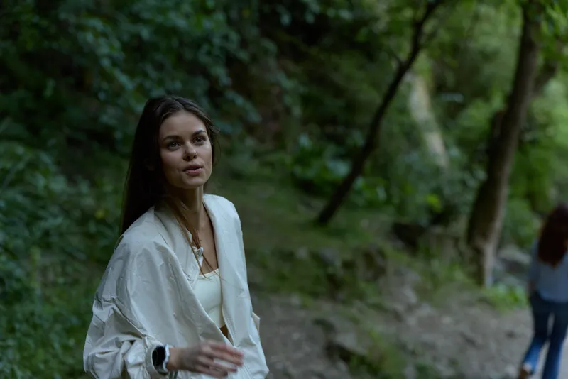 young woman in white jacket walking through lush green forest, natural light highlighting her thoughtful expression and serene outdoor atmosphere
