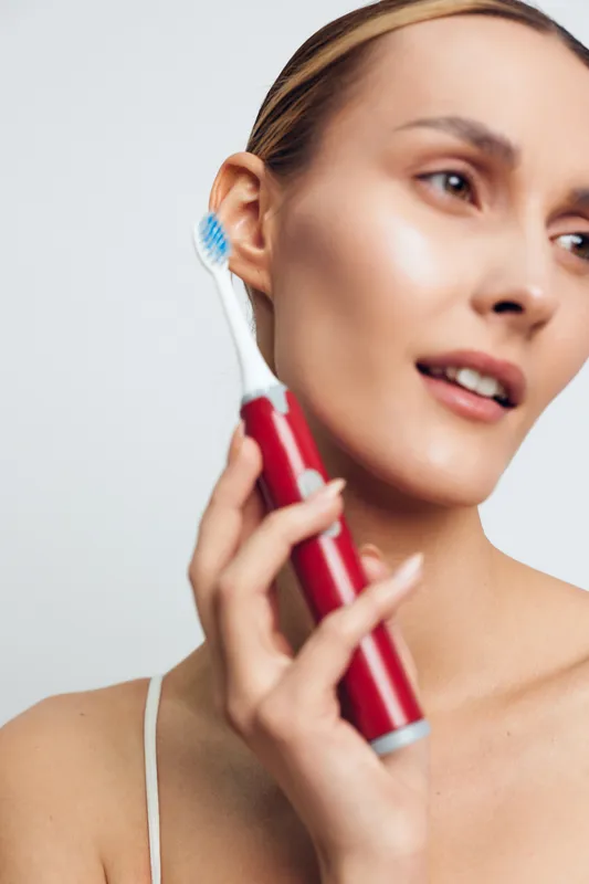 A young woman with clear skin holds a red electric toothbrush near her face, preparing for oral hygiene in a bright minimalistic setting. Concept for dental care and beauty routine.
