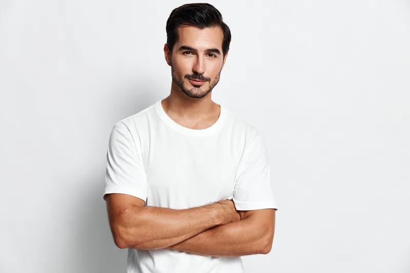 Confident young man in a white t-shirt poses with arms crossed, looking directly at the camera against a solid light background. Stylish casual portrait.