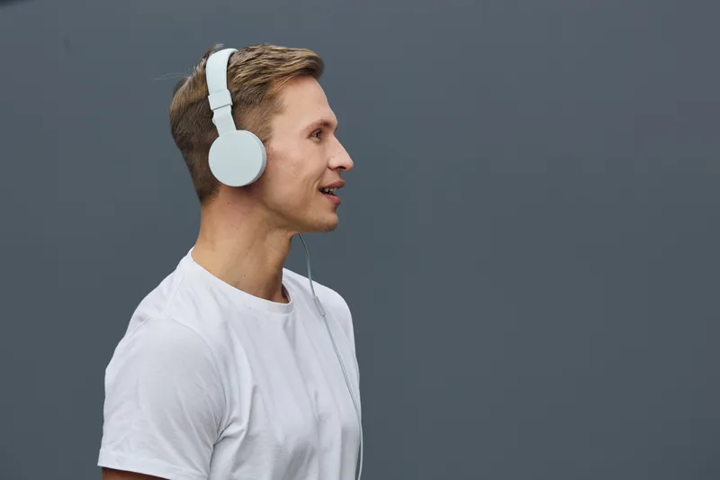 Young man with short brown hair wearing white t-shirt and large white headphones listening to music against a plain dark background. Casual style, person enjoying audio entertainment, studio shot, lif