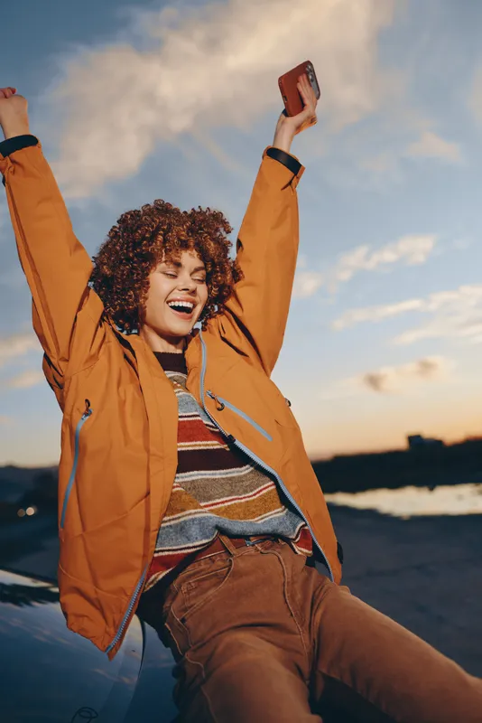 Woman lifestyle portrait outdoors at sunset, curly hair smiling with arms raised, orange jacket and headphones, warm evening sky and sea horizon, joyful relaxation, free feeling