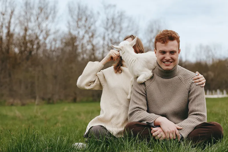 A couple enjoys an outdoor moment with a playful goat kid, sharing affection and laughter, sitting on grass in a park, showcasing a casual lifestyle and warm bond