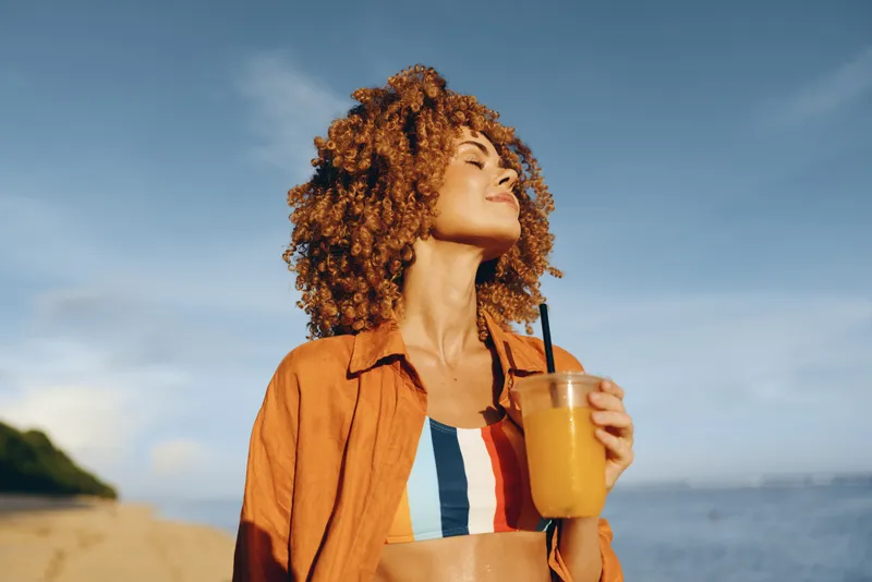 Young woman with curly red hair enjoys a refreshing drink on a sunny beach while wearing a colorful striped top and an open orange shirt.