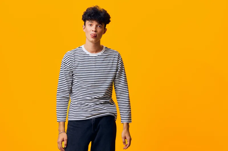 young man in striped shirt making playful face with tongue out against bright yellow background, casual expression, curly hair, modern style