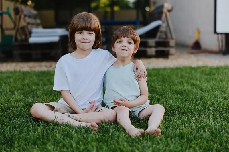 Two adorable boys sitting on the grass, smiling joyfully. The scene captures the essence of childhood fun, innocence, and friendship in a sunny environment.