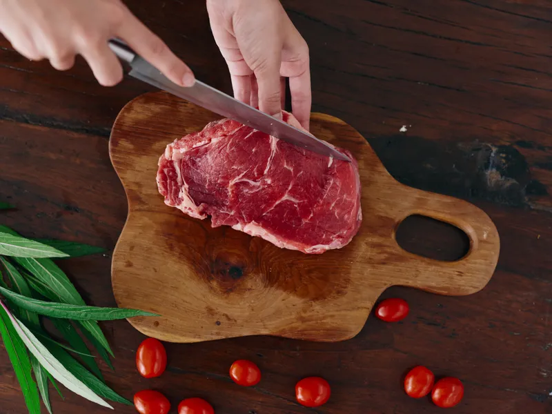 Женщина с ножом в руке нарезает свежее мясо стейка для жарки на кухне, на столе соль, перец и другие специи, красные помидоры черри и зелень, приготовление ужина в домашней кухне