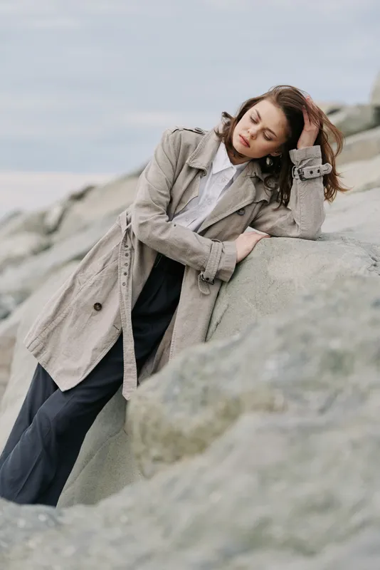 Young woman in beige coat leans on large rock outdoors, eyes closed and hand in hair. Natural light and muted colors create calm, introspective mood in coastal landscape setting.