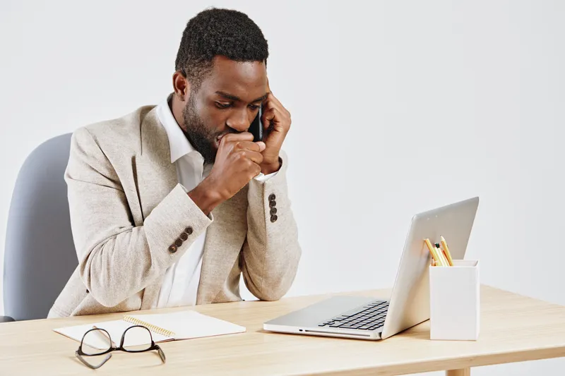 Professional business man talking on mobile phone while working at his desk, using laptop, with glasses and notepad, in modern office environment. People lifestyle concept, office communication, techn