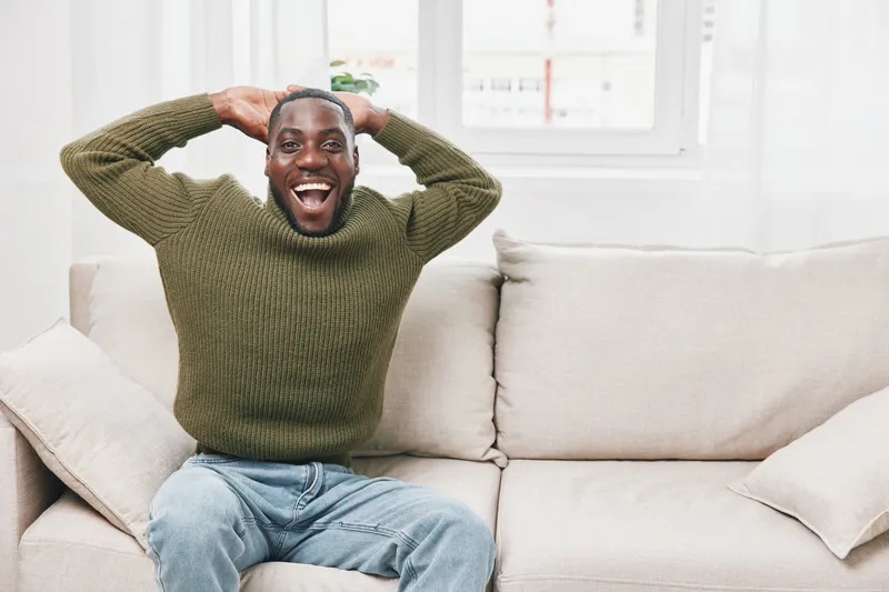 Happy young African American man in cozy green sweater celebrating on a light sofa indoors, expressing joy and excitement in a bright, modern living room