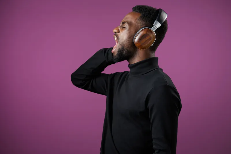 Young African man passionately singing into headphones against a vibrant purple background, showcasing emotion and music enjoyment