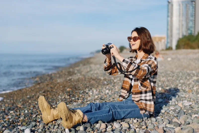 Woman photographer sits on a pebble beach with a camera, capturing seaside scenery in casual outdoor style. Sunny day, plaid shirt, jeans, boots, coastal backdrop, relaxed creative moment