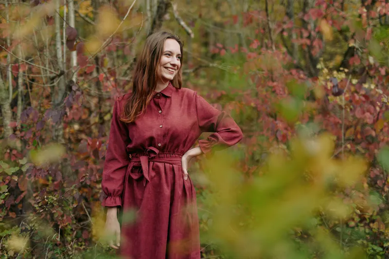 A woman in a burgundy dress stands in a sunlit autumn garden, conveying authenticity and credibility through a natural pose, relaxed smile, and authentic surroundings.