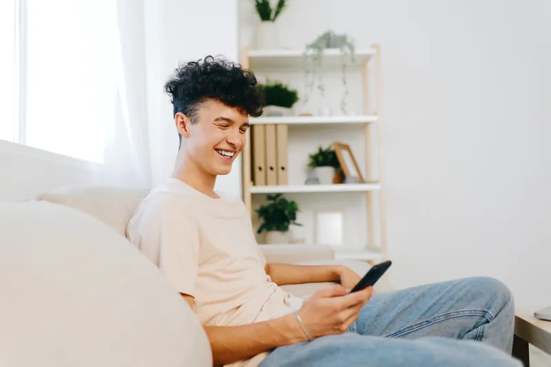 Улыбающийся молодой человек сидит на светлом диване и пользуется смартфоном в светлой современной комнате