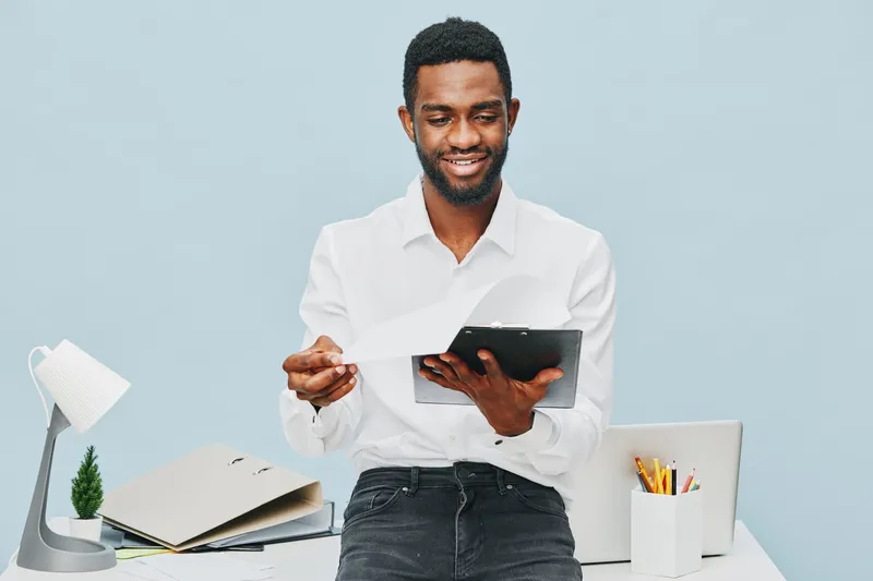 Smiling man in white shirt holding tablet device while working at modern office desk with stationery and laptop, professional corporate environment, confident business person, indoor studio shot.
