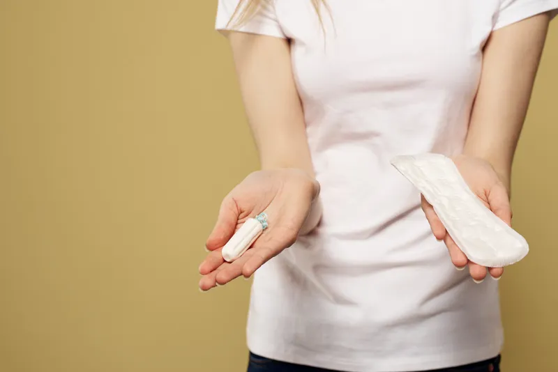 Девушка держит в руке тампон и прокладку на бежевом фоне, на ней белая футболка, кадр обрезан