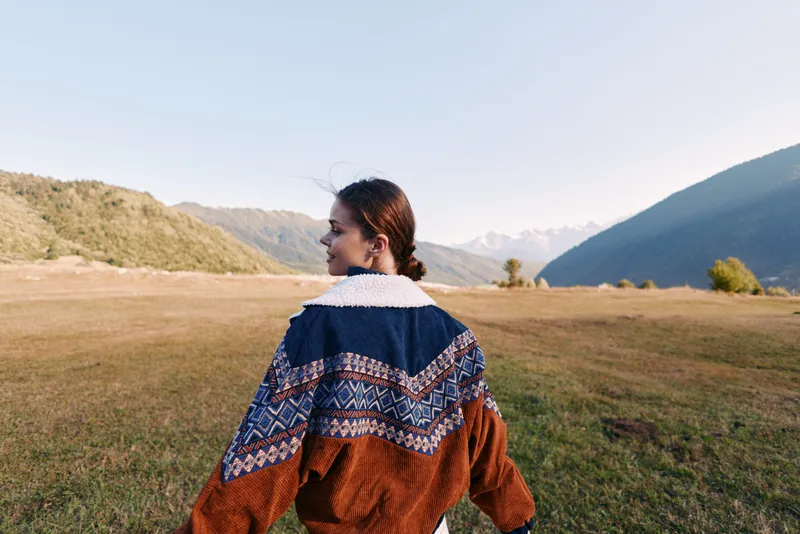 Woman in sweater walks across a meadow and landscape with mountains in the distance, autumn light and travel vibe, nature scene showing backview portrait and peaceful outdoors journey.