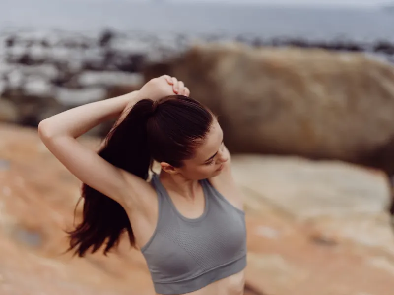 Young woman stretching outdoors on a rocky beach, wearing a gray sports bra and enjoying a moment of relaxation, reflecting fitness and well being