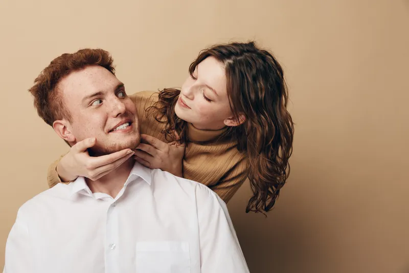 Young couple in casual clothes share a tender moment, woman gently holding man’s face. Warm beige background creates intimate and loving atmosphere, concept of affection and relationship.