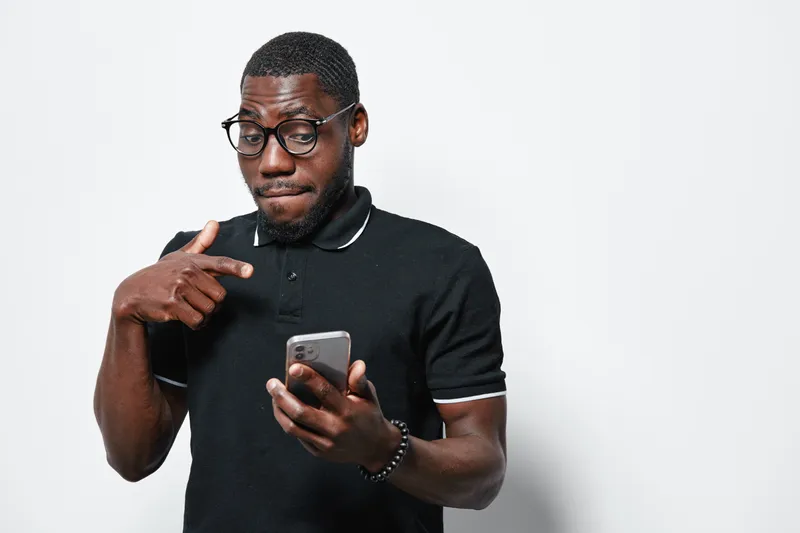 African American man with dark skin wearing glasses and black polo shirt pointing at smartphone with thoughtful expression on white background. Technology, communication and casual style.