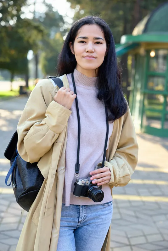 Photographer in a beige coat with camera hanging at the neck, standing along a sunlit park path, outdoor casual shoot with street style and urban exploration vibes