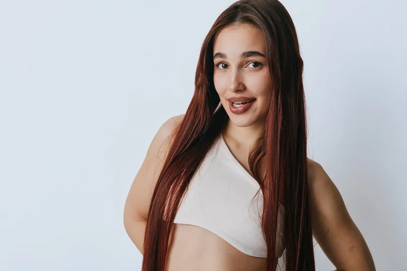 Young woman with long red hair wearing a white crop top posing with hands behind back on white background. Portrait of confident female model with natural makeup and relaxed expression.