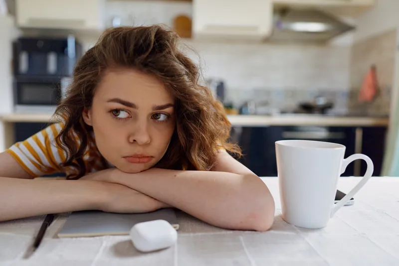 A young woman with wavy hair rests her head on crossed arms on a bright kitchen table, a white mug and a computer mouse nearby, conveying fatigue and boredom in a candid home moment.