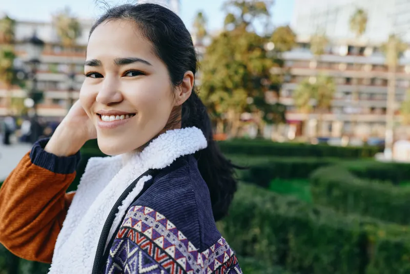 Smiling woman outdoors wearing a patterned knit sweater and scarf in a park setting with greenery and distant buildings, casual fashion mood and a bright friendly atmosphere