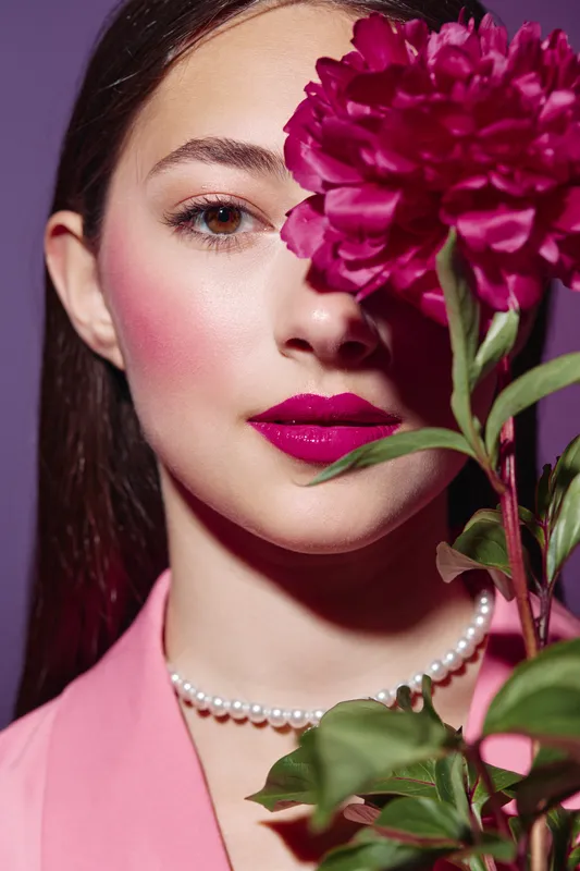 Young woman with bright pink lips and blush holding a big magenta flower near her face. Close-up fashion beauty portrait on lavender background. Concept of color style.