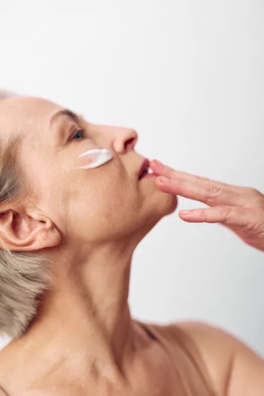 Mature woman applies white cream on face with fingers in soft natural light. Skincare concept for aging skin care, beauty routine, and self-care in minimalistic studio setting.