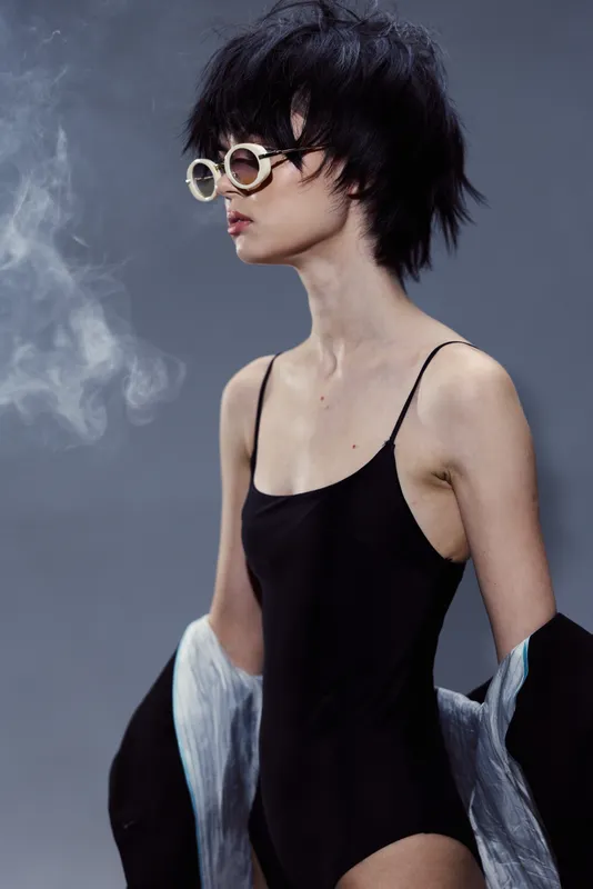 Stylish woman with short hair wearing sunglasses and black bodysuit with smoky background and blue jacket draped on arms