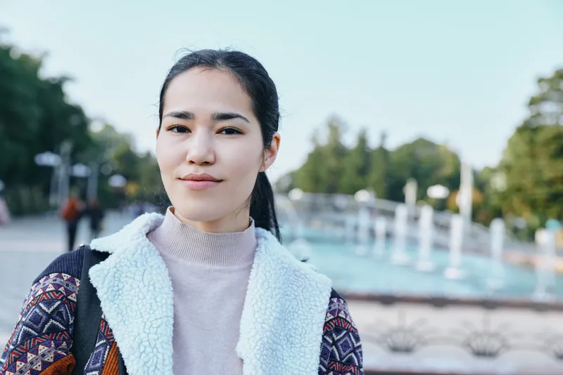 Woman standing outdoors in a park beside a decorative fountain, wearing a soft sweater and patterned jacket, calm expression, natural light, casual urban lifestyle shot.