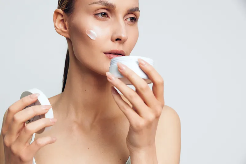 Young woman applying white face cream on cheek holding open jar in hand. Skincare routine concept with natural skin and clear background. Soft lighting highlights healthy skin texture.