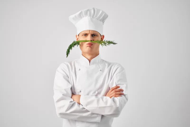 Повар держит кухонную утварь рядом с лицом, на фоне зелени, в ресторане