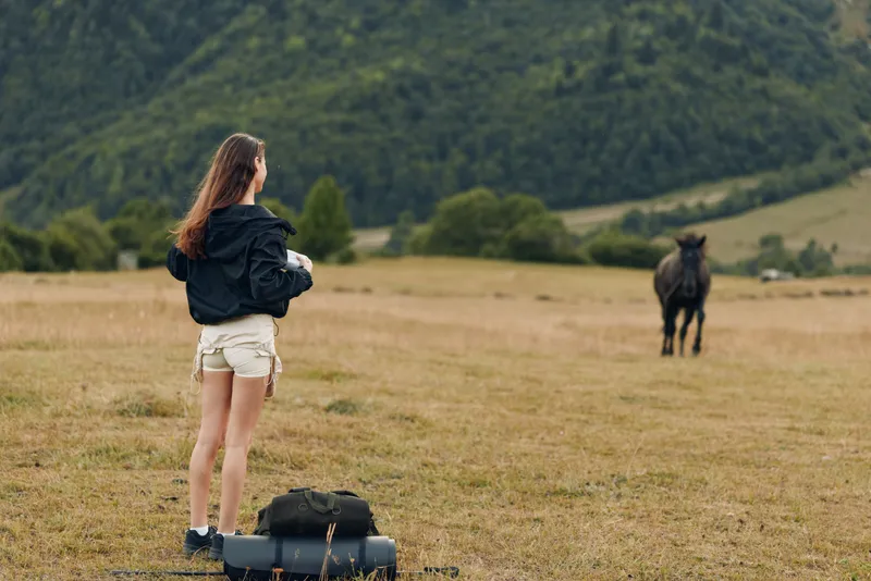 A young woman stands in an open field, gazing at a dark horse in the distance, with a backpack and luggage at her feet, capturing a calm rural moment.