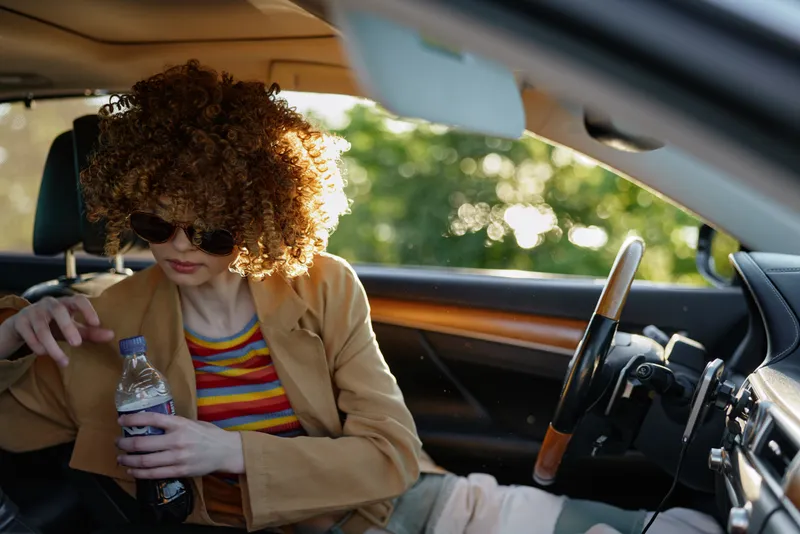 Curly haired woman sits in a car interior wearing a striped shirt and tan blazer, holding a water bottle while soft daylight filters through the window, creating a calm, casual mood.