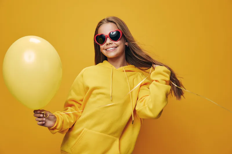 Smiling girl wearing red heart sunglasses and yellow hoodie holding a yellow balloon on yellow background with playful attitude and casual style.
