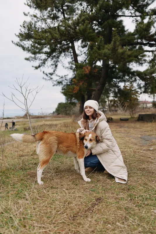 Woman in a warm coat hugs a friendly dog in a park, autumn leaves and tall pines under an overcast sky, a tender outdoor moment of companionship.